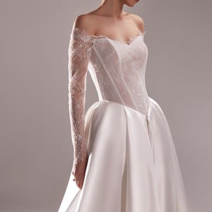 K&ouml;nnte beinhalten: Elegantes wei&szlig;es Hochzeitskleid mit schulterfreiem Ausschnitt und langen Spitzen&auml;rmeln. Das Oberteil ist mit zarter Spitze verziert, und der Rock flie&szlig;t anmutig. Ein klassisches Brautkleid f&uuml;r einen besonderen Anlass.