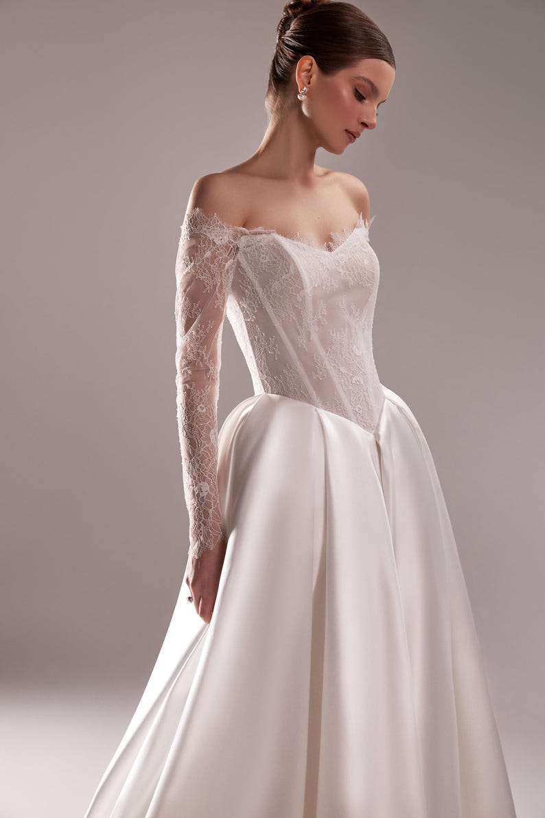K&ouml;nnte beinhalten: Elegantes wei&szlig;es Hochzeitskleid mit schulterfreiem Ausschnitt und langen Spitzen&auml;rmeln. Das Oberteil ist mit zarter Spitze verziert, und der Rock flie&szlig;t anmutig. Ein klassisches Brautkleid f&uuml;r einen besonderen Anlass.