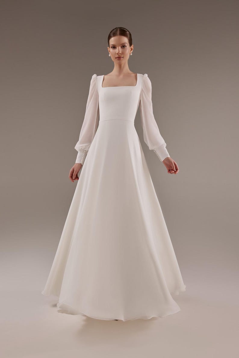 May include: A white wedding dress with a square neckline and long sleeves. The dress has a fitted bodice and a flowing skirt.
