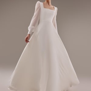 May include: A white wedding dress with long sleeves and a square neckline. The dress has a fitted bodice and a flowing skirt.