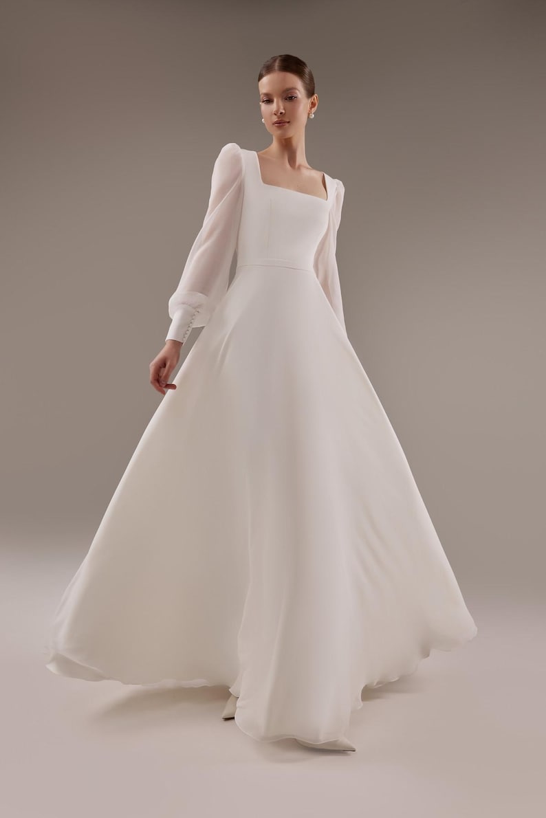 May include: A white wedding dress with long sleeves and a square neckline. The dress has a fitted bodice and a flowing skirt.