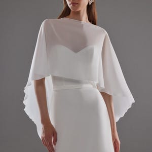 May include: A white bridal gown with a sweetheart neckline and a sheer, flowing cape. The dress has a fitted bodice and a streamlined skirt. The cape drapes over the shoulders, creating a soft, elegant silhouette. The model is wearing pearl earrings.