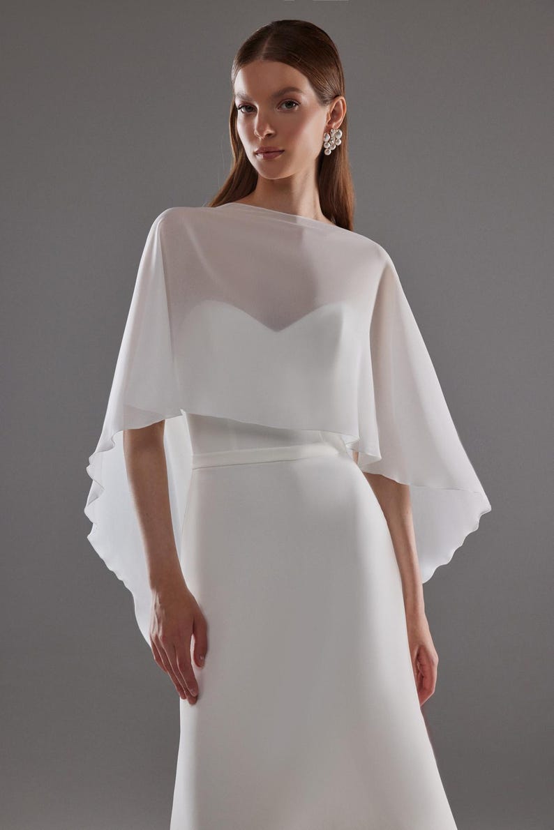 May include: A white bridal gown with a sweetheart neckline and a sheer, flowing cape. The dress has a fitted bodice and a streamlined skirt. The cape drapes over the shoulders, creating a soft, elegant silhouette. The model is wearing pearl earrings.