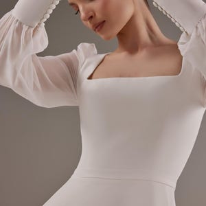 May include: A white wedding dress with a square neckline and long sleeves. The sleeves are made of sheer fabric with a gathered detail at the shoulder. The dress has a fitted bodice and a flowing skirt.
