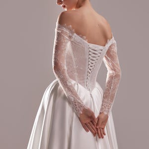 K&ouml;nnte beinhalten: Wei&szlig;es Satin-Brautkleid mit Spitzenoberteil und langen Spitzen&auml;rmeln. Der R&uuml;cken des Kleides hat einen Korsettverschluss. Die H&auml;nde der Braut sind vor ihr gefaltet.
