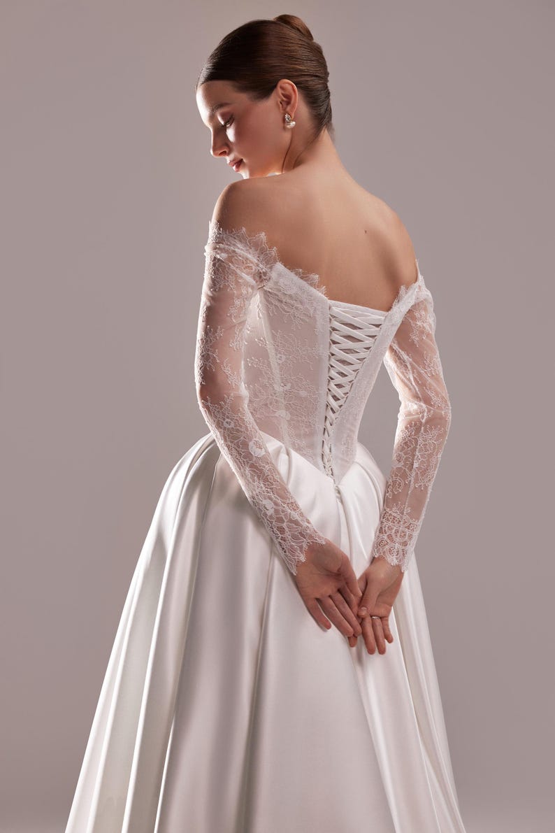 K&ouml;nnte beinhalten: Wei&szlig;es Satin-Brautkleid mit Spitzenoberteil und langen Spitzen&auml;rmeln. Der R&uuml;cken des Kleides hat einen Korsettverschluss. Die H&auml;nde der Braut sind vor ihr gefaltet.