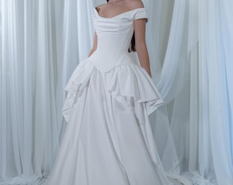 Könnte beinhalten: Ein weißes schulterfreies Hochzeitskleid mit einem figurbetonten Oberteil und einem weiten Rock. Das Kleid hat ein mehrschichtiges Design und eine fließende Silhouette. Das Model steht vor weißen Vorhängen.