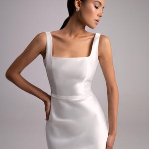 May include: A white sleeveless dress with a square neckline and a fitted silhouette. The dress has a simple design and a smooth, satin-like texture.