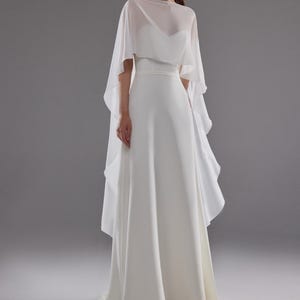 May include: A white wedding dress with a flowing skirt and a sheer, cape-like overlay. The dress features a fitted bodice and a simple, elegant design. The model is wearing earrings and has her hair pulled back.