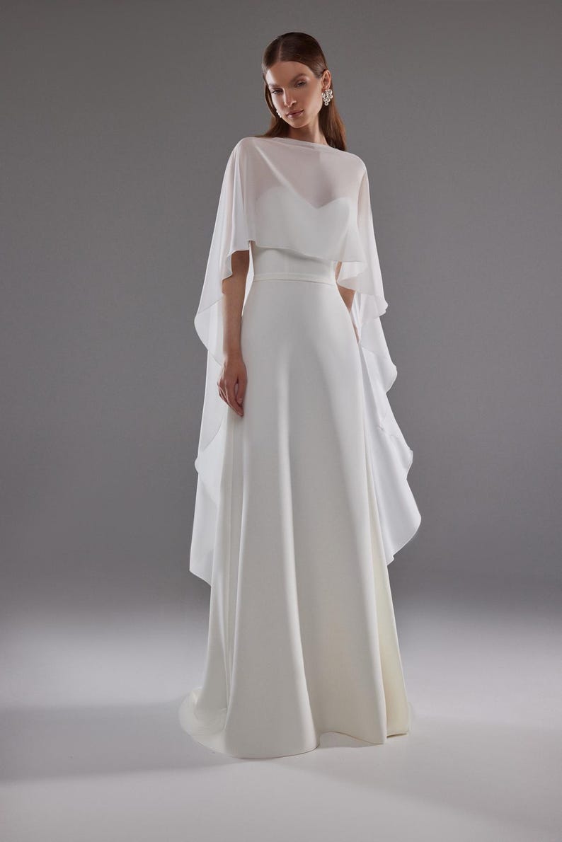 May include: A white wedding dress with a flowing skirt and a sheer, cape-like overlay. The dress features a fitted bodice and a simple, elegant design. The model is wearing earrings and has her hair pulled back.