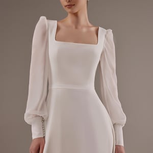 May include: A white wedding dress with long sleeves and a square neckline. The sleeves are sheer and have a gathered detail at the cuff. The dress has a fitted bodice and a flowing skirt.