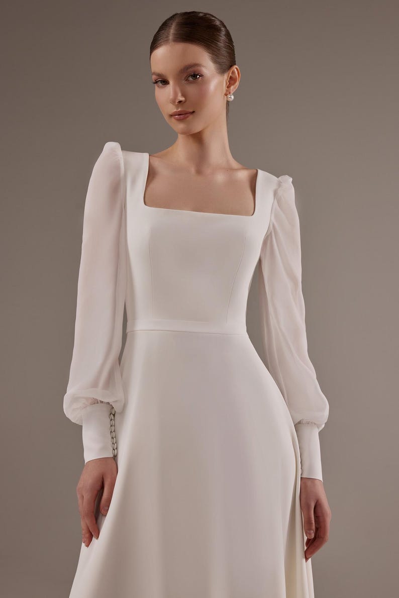 May include: A white wedding dress with long sleeves and a square neckline. The sleeves are sheer and have a gathered detail at the cuff. The dress has a fitted bodice and a flowing skirt.