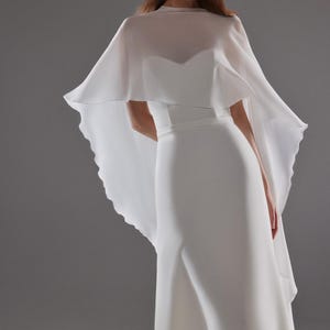 May include: A white wedding dress with a flowing cape. The dress has a fitted bodice and a flared skirt. The cape is sheer and drapes over the shoulders. The model is wearing earrings.