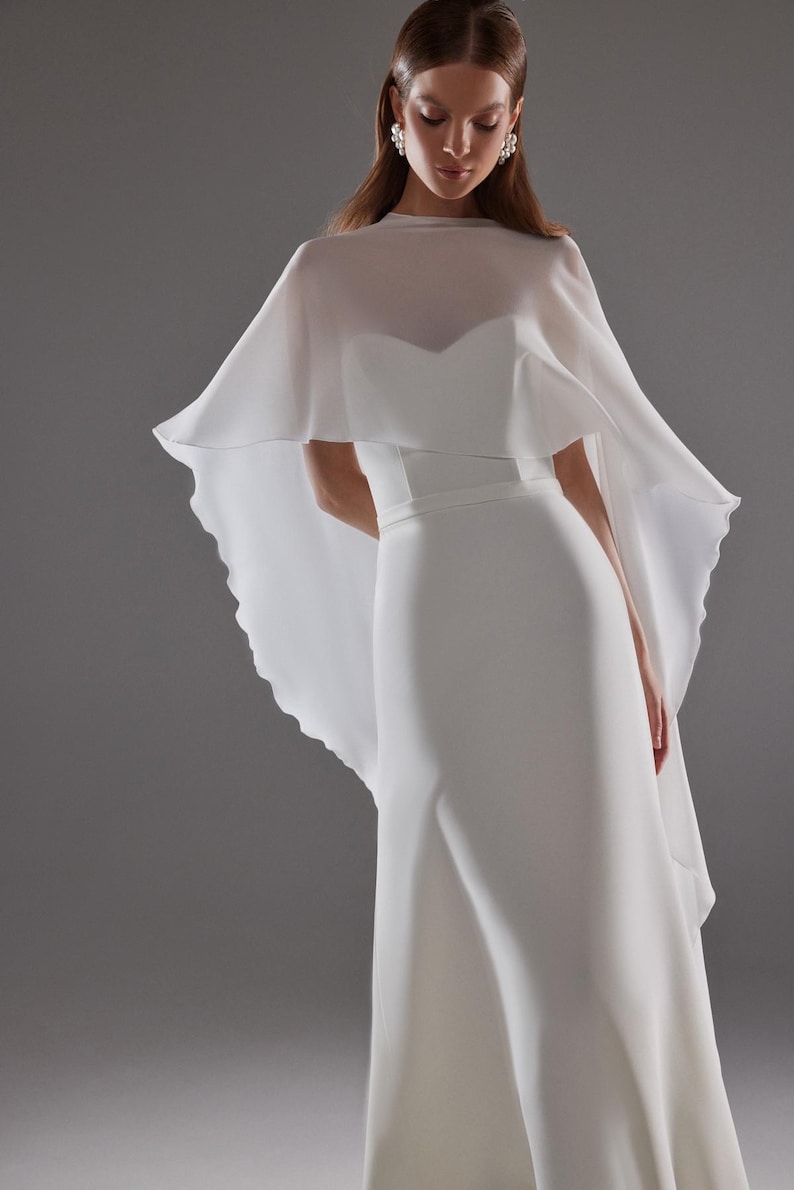 May include: A white wedding dress with a flowing cape. The dress has a fitted bodice and a flared skirt. The cape is sheer and drapes over the shoulders. The model is wearing earrings.