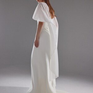 May include: A white bridal gown with a flowing skirt and a sheer, cape-like overlay. The dress has a simple, elegant design, with a slight train. The model is wearing earrings and has long, light brown hair.