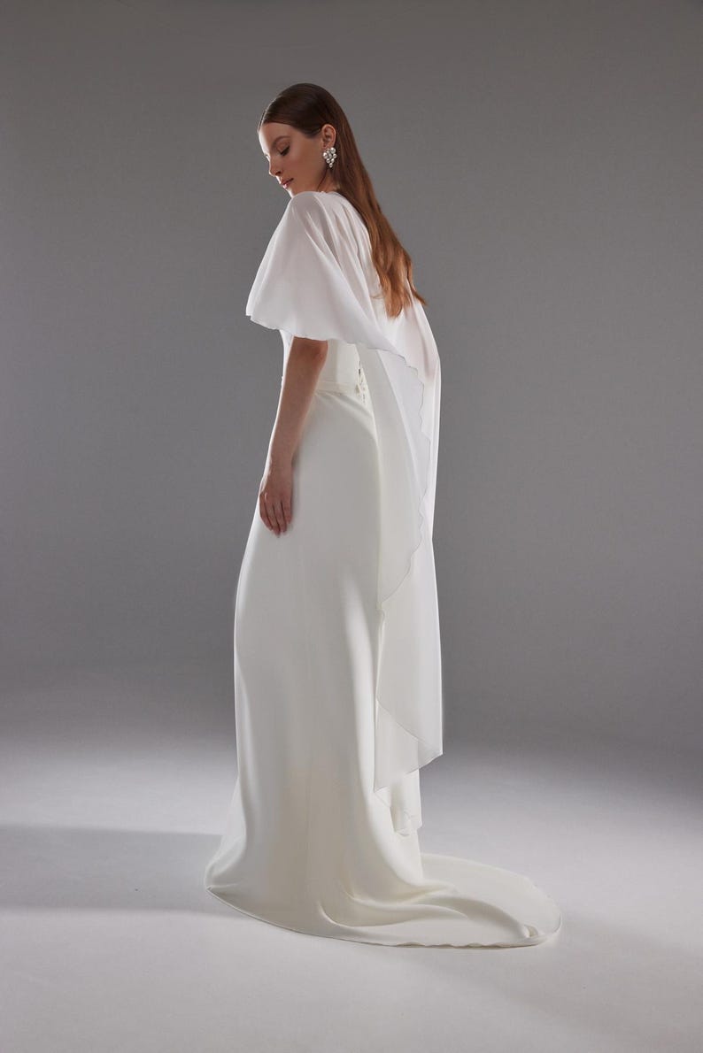 May include: A white bridal gown with a flowing skirt and a sheer, cape-like overlay. The dress has a simple, elegant design, with a slight train. The model is wearing earrings and has long, light brown hair.