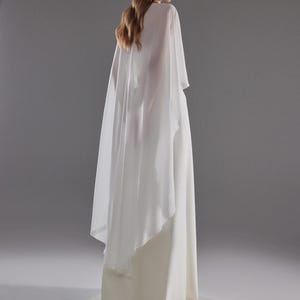 May include: A white bridal gown with a flowing skirt and a sheer cape. The dress has a simple, elegant design, perfect for a wedding. The model is facing away from the camera, showcasing the back of the dress and cape against a gray background.