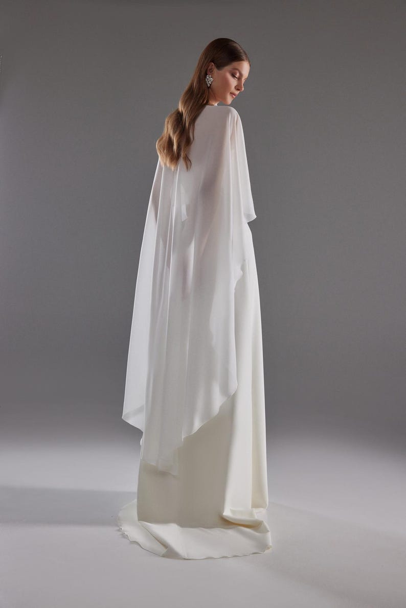 May include: A white bridal gown with a flowing skirt and a sheer cape. The dress has a simple, elegant design, perfect for a wedding. The model is facing away from the camera, showcasing the back of the dress and cape against a gray background.