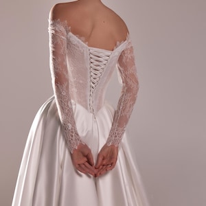 K&ouml;nnte beinhalten: Wei&szlig;es Hochzeitskleid mit Satinrock und Spitzenoberteil. Die schulterfreien &Auml;rmel sind lang und aus Spitze. Der R&uuml;cken des Kleides hat einen Korsettverschluss.