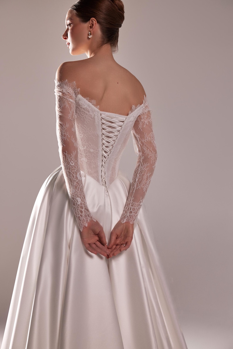 K&ouml;nnte beinhalten: Wei&szlig;es Hochzeitskleid mit Satinrock und Spitzenoberteil. Die schulterfreien &Auml;rmel sind lang und aus Spitze. Der R&uuml;cken des Kleides hat einen Korsettverschluss.
