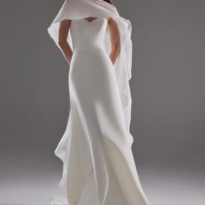 May include: A white, floor-length wedding gown with a sweetheart neckline and a flowing cape. The dress has a fitted bodice and a flared skirt. The cape drapes over the shoulders and arms. The model is wearing white heels.
