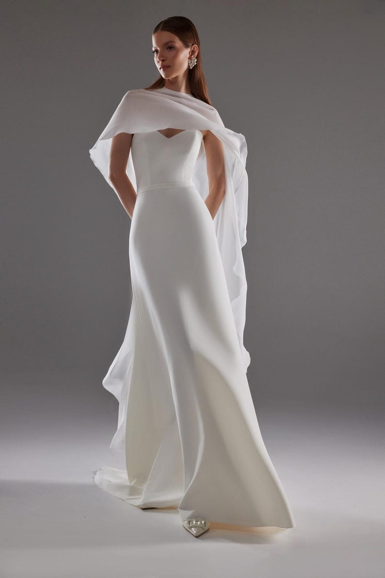 May include: A white, floor-length wedding gown with a sweetheart neckline and a flowing cape. The dress has a fitted bodice and a flared skirt. The cape drapes over the shoulders and arms. The model is wearing white heels.
