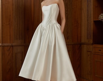 May include: A strapless, white satin dress with a fitted bodice and a full, knee-length skirt. The dress features a structured design and is suitable for formal events. The model is wearing white heels. The background is a wooden wall and floor.