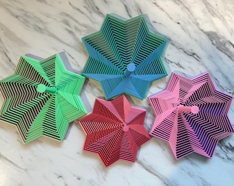 May include: A collection of five star-shaped, layered objects in shades of green, blue, red, pink, and purple. Each has a central peg. The objects are arranged on a marble-patterned surface. The design features a series of concentric lines.