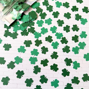 May include: Green shamrock confetti scattered on a white brick background. A clear plastic bag filled with more shamrock confetti is in the upper left corner. The confetti is in various shades of green.