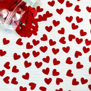 Puede incluir: Una colecci&oacute;n de peque&ntilde;os confetis planos en forma de coraz&oacute;n rojo esparcidos sobre una superficie blanca con patr&oacute;n de ladrillo. Una bolsa de pl&aacute;stico transparente e iridiscente, parcialmente abierta, se sit&uacute;a en la esquina superior izquierda.