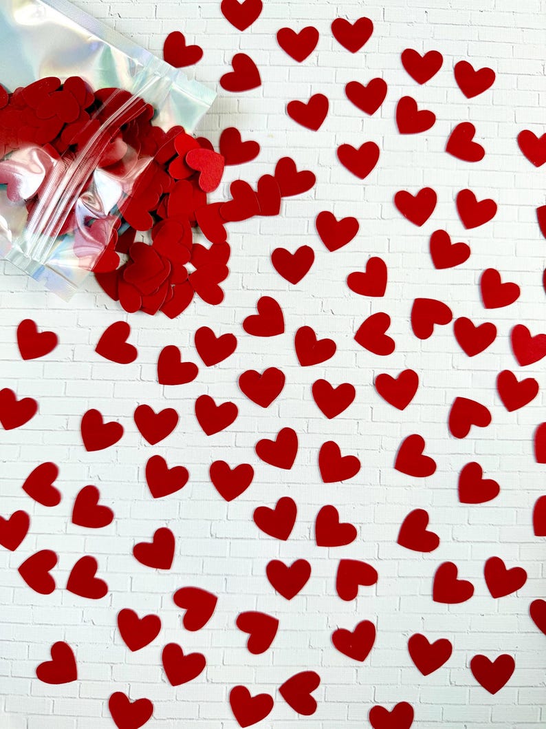 Puede incluir: Una colecci&oacute;n de peque&ntilde;os confetis planos en forma de coraz&oacute;n rojo esparcidos sobre una superficie blanca con patr&oacute;n de ladrillo. Una bolsa de pl&aacute;stico transparente e iridiscente, parcialmente abierta, se sit&uacute;a en la esquina superior izquierda.