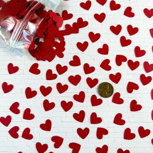 Puede incluir: Una colecci&oacute;n de peque&ntilde;os recortes planos en forma de coraz&oacute;n de color rojo esparcidos sobre una superficie blanca, con una parte de los corazones contenida en una bolsa de pl&aacute;stico iridiscente transparente. Una moneda de cobre sirve de escala.