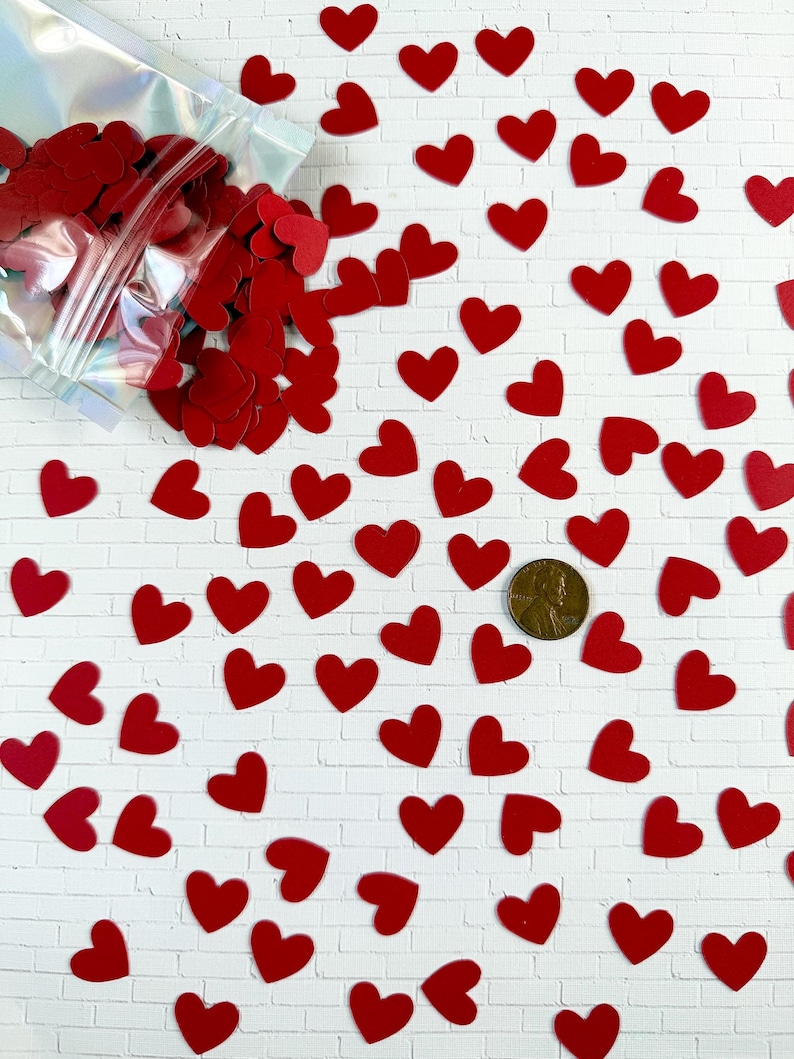 Puede incluir: Una colecci&oacute;n de peque&ntilde;os recortes planos en forma de coraz&oacute;n de color rojo esparcidos sobre una superficie blanca, con una parte de los corazones contenida en una bolsa de pl&aacute;stico iridiscente transparente. Una moneda de cobre sirve de escala.