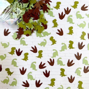 May include: A collection of dinosaur-themed confetti in shades of green and brown, including dinosaur shapes and footprints. The confetti is scattered on a white brick background, with some spilling from a clear bag.