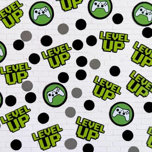May include: Confetti with a gaming theme. The confetti includes green and white controller icons in green circles, the words "LEVEL UP" in green pixelated font, and black and gray circular confetti pieces. The background is a white brick wall.