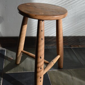 May include: A handcrafted wooden stool with a round seat and three angled legs. The stool is made of light brown wood, with visible wood grain and dark accents. The design is simple and rustic, suitable for various interior styles.