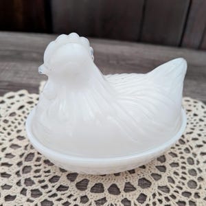 Puede incluir: Un plato blanco en forma de gallina con tapa. El plato es de vidrio opaco y se asienta sobre un tapete de encaje. La gallina tiene plumas detalladas y una cresta. Un encantador artículo de cocina vintage.