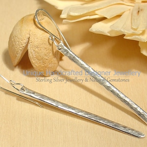 Peut inclure: Une paire de longues boucles d'oreilles pendantes fines et argentées avec une finition martelée et texturée. Chaque boucle présente une fine tige droite suspendue à un crochet incurvé. La surface métallique reflète la lumière avec éclat.