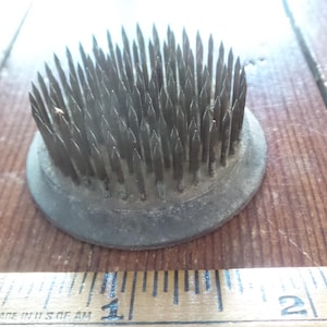 Op de afbeelding: Een vintage bloemenkikker, een ronde, donkergrijze metalen basis met talrijke scherpe, omhoog wijzende metalen pinnen. De bloemschikgereedschap ligt op een houten oppervlak.