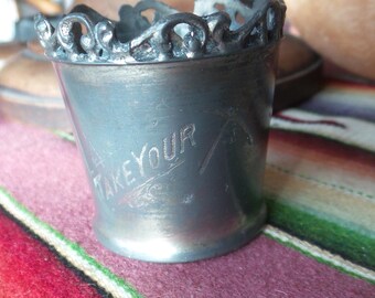 May include: A silver cup with an ornate rim and the words "Take Your" engraved on the side. The cup is sitting on a colorful striped blanket.