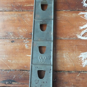 Könnte beinhalten: Ein vintage Eierhalter aus Metall mit sechs Fächern und einem Griff. Der Halter ist dunkelgrau lackiert und hat die Buchstaben "SS" und einen Kreis mit einem "C" darin auf der Vorderseite.