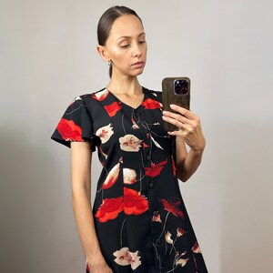 Peut inclure: Une femme portant une robe noire &agrave; imprim&eacute; floral rouge et blanc. La robe a un devant boutonn&eacute; et des manches courtes. Elle tient un smartphone dans sa main droite.