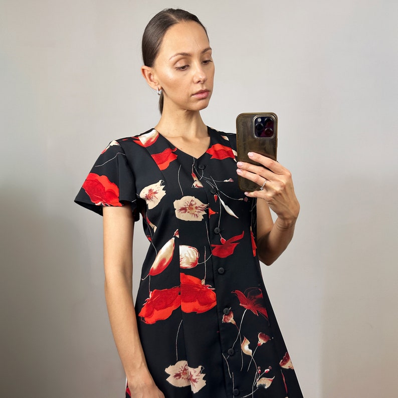 Peut inclure: Une femme portant une robe noire &agrave; imprim&eacute; floral rouge et blanc. La robe a un devant boutonn&eacute; et des manches courtes. Elle tient un smartphone dans sa main droite.