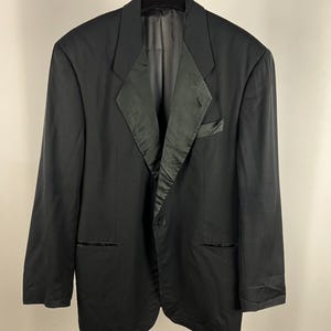 May include: A black blazer with a satin lapel and a single-button closure. The blazer has two front pockets and is hanging on a black hanger. The brand name "roberto cavalli" is visible on the hanger.