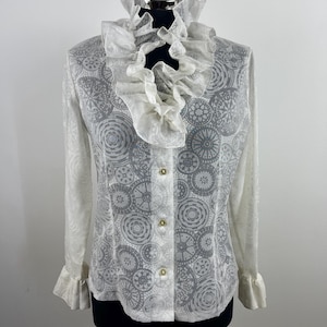 May include: A white blouse with a high ruffled collar and long sleeves. The blouse features a pattern of gray circular designs and is buttoned down the front with gold-colored buttons. The sleeves have ruffled cuffs.