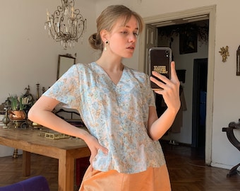 May include: A woman wearing a light blue and white floral print blouse with a button-up front and a peach-colored skirt. She is holding a black phone case with gold lettering that says "Stay Weird, Stay Kind, Stay True".