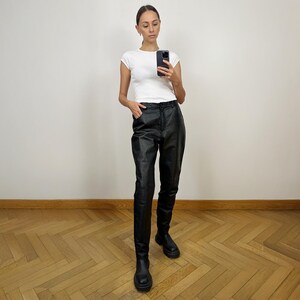 Peut inclure: Une femme portant un t-shirt blanc et un pantalon en cuir noir. Elle est debout sur un sol en bois et regarde son t&eacute;l&eacute;phone.
