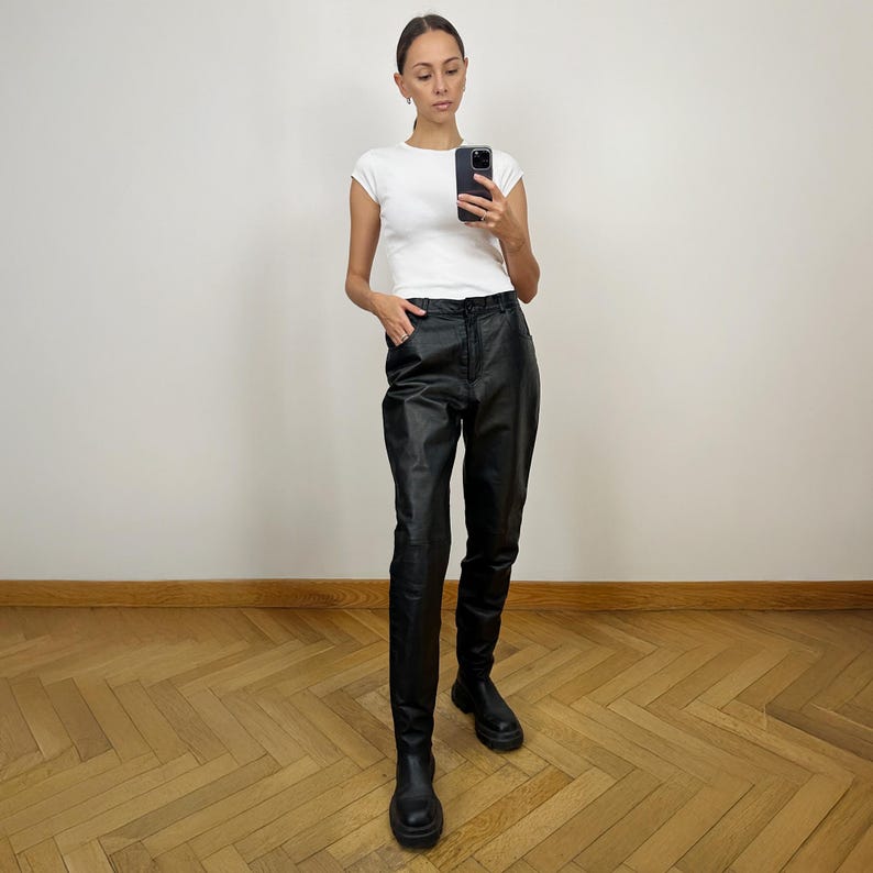 Peut inclure: Une femme portant un t-shirt blanc et un pantalon en cuir noir. Elle est debout sur un sol en bois et regarde son t&eacute;l&eacute;phone.