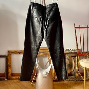 Peut inclure: Pantalon en cuir noir suspendu &agrave; un cintre. Le pantalon a une jambe droite et une fermeture &eacute;clair. Le pantalon est suspendu devant un mur blanc.