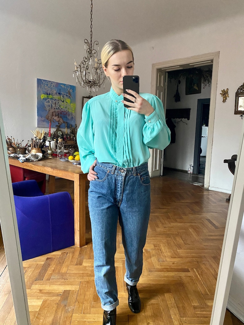 May include: A light turquoise blouse with a high collar and pleated front, paired with blue denim jeans. The blouse has long, puffed sleeves. The jeans are cuffed at the ankles. Black boots complete the outfit.